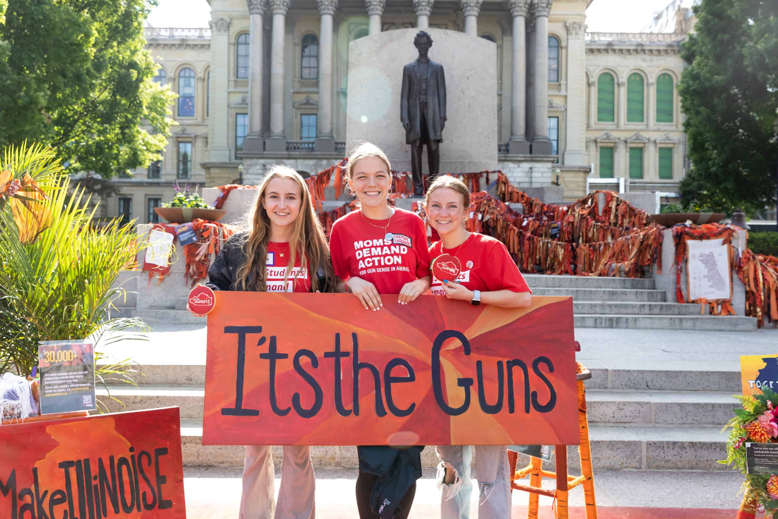 Students holding an Arts4Impact 'It's the Guns' sign in front of the IL State Capitol.